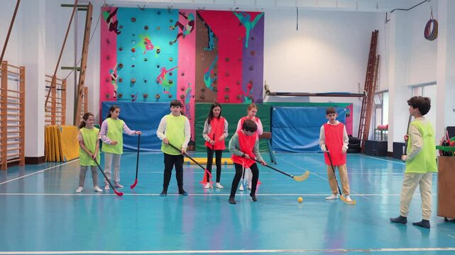 Energetic students wearing vests learning and practicing floorball together during physical education class in bright school gymnasium, demonstrating teamwork and athletic skills