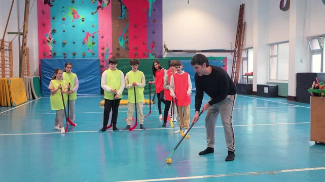 Floorball coach demonstrating proper stick handling and ball control techniques to a group of young students in a gym