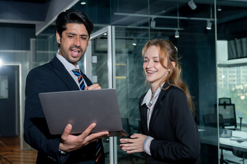 Male and female business colleagues standing side by side looking at laptop screen in office with surprised facial expressions while discussing online data or presentation together in modern workspace
