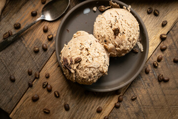 Two scoops of gourmet coffee flavored homemade ice cream on dark brown shallow bowl, wooden table, with coffee beans scattered around.