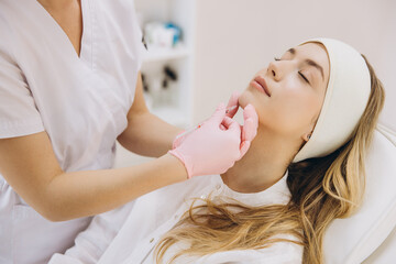 Beautician performing chin augmentation procedure on young woman lying on medical chair, enhancing...