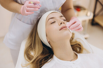 Cosmetologist cleaning face skin of young blond woman with cotton pad, preparing for injection...