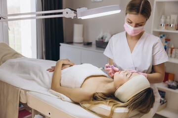 Obraz premium Beautician wearing mask and gloves injecting filler into lips of young woman lying on bed in cosmetology clinic with magnifying lamp