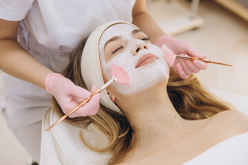 Professional cosmetologist applying facial mask with soft brushes, carefully treating client's skin for deep cleansing and relaxation