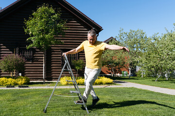 Senior man carefully stepping off a metal garden ladder on a sunny day in front of a wooden house. 