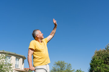 Senior man standing outdoors on a sunny day, raising his hand and looking up with a smile. Concept of a positive and active lifestyle, gardening, wellness, aging well, or retirement planning.