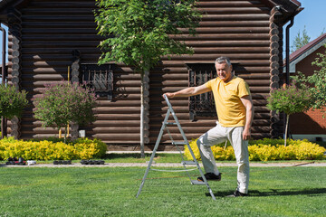 Senior man in casual clothes standing confidently on a garden ladder near a wooden log house.