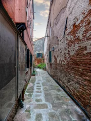 A narrow Venetian alleyway framed by two buildings, one with a weathered red facade, the other showcasing exposed bricks. Travel in Italy.