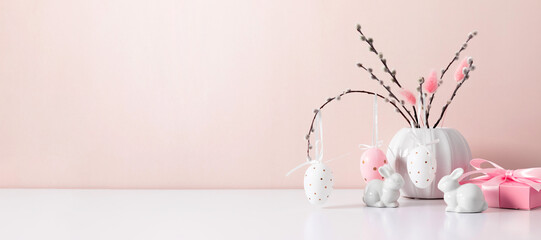 Obraz premium Cozy Easter composition on a white shelf against the background of a wall, a blooming willow branch with Easter eggs, a gift and a cute bunny. Easter background, banner.
