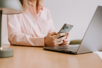 Woman uses smartphone while sitting at desk with laptop and lamp in a modern workspace