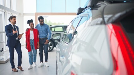 Attractive manager offering to get acquainted with new cars on tablet device. Couple listening with interest and reviewing information on gadget. Buyers thinking about buying car in showroom.