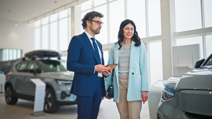 Obraz premium Beautiful Caucasian woman asking questions to auto dealer. People communicating together while standing in auto salon. Handsome man in formal suit holding tablet device while talking with client.