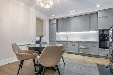 Kitchen interior with modern design. Beautiful ceiling chandelier over the dining table made of dark wood, light gray kitchen set.