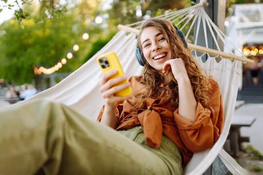 Beautiful woman relaxing in hammock by the lake wearing wireless headphones and using phone. Young woman enjoying sunny weather in park. Concept of relaxation, fun, blogging.