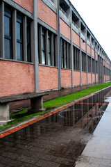 A contemporary brick building with rows of windows is reflected in water puddles after the rain, creating a balanced urban scene