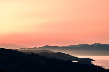 Mountains silhouetted against a coastline bathed in pink and orange hues at dusk. The horizon fades into a soft mist, creating a serene atmosphere.