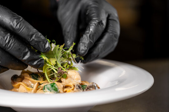 A chef's hands in black gloves garnish a plate of creamy pasta with fresh microgreens, emphasizing culinary craftsmanship and presentation in fine dining
