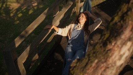 View from above of beautiful Caucasian girl laying in hammock in middle of garden. Swinging slowly while resting. Enjoying sunset after hard working day at farm. Resting in evening. Relaxation.