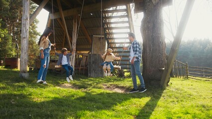 Caring Caucasian parents pushing their children on swing. Little boy and girl loving activity with mother and father. Family spending time together on sunny day at farm. Tall trees in background.