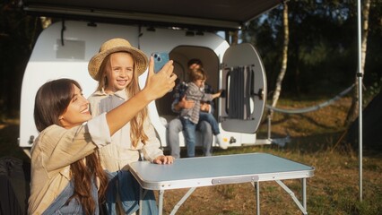 Obraz premium Caring Caucasian mother holding phone with one hand while trying to take selfie photo with beautiful daughter. Dad in background playing with their son. Camping together as family in RV.