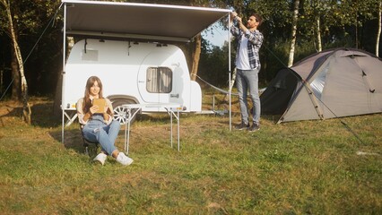 Caucasian family having relaxing free time in nature. Woman sitting in front on vehicle on small chair and using tablet device. Man setting up camp. Family holiday. Weekend together.