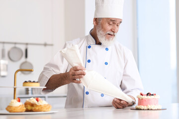 Elderly confectioner with pastry bag making cake at table in kitchen