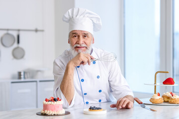 Elderly pastry chef holding whisk at table with desserts in kitchen