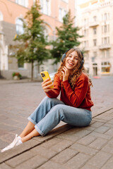 A cute woman in wireless headphones and with a phone enjoys a walk along the sunset streets of the city. Beautiful tourist listening to music on headphones outdoors. The concept of relaxation.