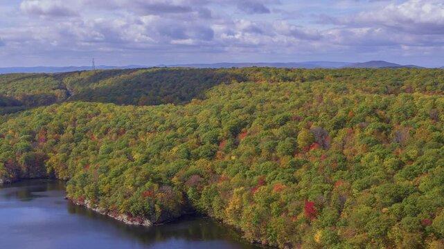 This 4K drone shot captures Canopus Lake in Kent, NY, with sunlight on the autumn forest beside the lake, creating a beautiful fall scene under cloudy weather.