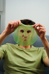 Relaxed man with hydrating face mask at home. Man is applying facial mask for rejuvenate...