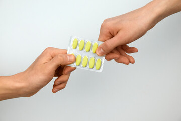 Two hands exchanging a blister pack of yellow pills against a neutral background.