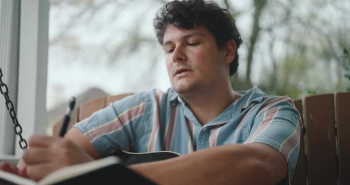 Young male musician sitting on a porch, playing guitar and writing a song