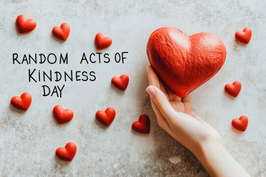 A hand holding a red heart with random acts of kindness day text and small red hearts scattered around it