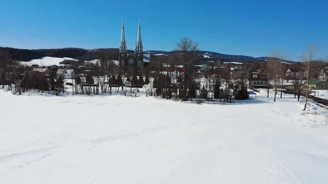 The village of Val-Brillant and its Saint-Pierre-du-Lac Church seen from Lake Matapedia in a mountainous setting whitened by winter snow under a cloudless blue sky.