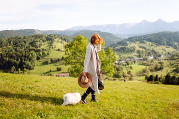 Naklejka premium Female tourist having fun with her small white dog on grassy hillside. Back view of beautiful woman with her pet enjoying mountain landscape at sunset. Freedom, travel and fun concept.