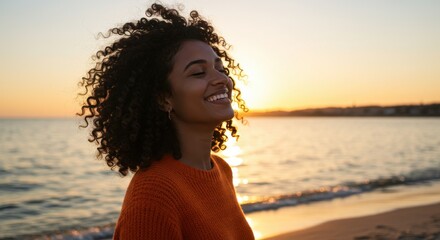 Woman with curly hair smiling at sunset on beach with orange sweater and ocean background view