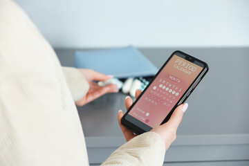 Menstruation. Woman holding smartphone with period calendar application and taking tampon indoors, closeup