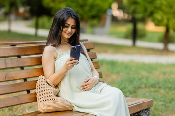 Fototapeta premium beautiful brunette pregnant woman, sitting on a bench in the park, holding a phone in her hands