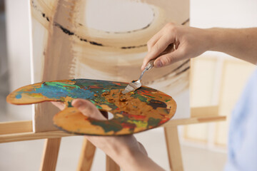 Young man mixing paints on palette near painting in artist's studio, closeup