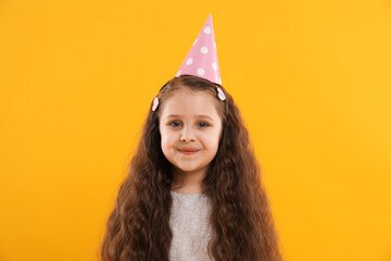 Cute little girl in conical paper hat on yellow background. Birthday surprise party