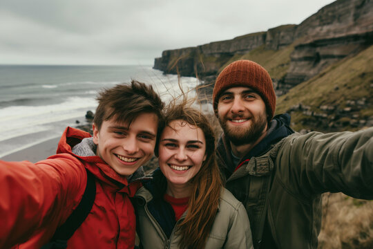 Three friends smiling and taking selfie on windswept cliffside overlooking ocean. Clouds and waves create serene, scenic backdrop with rugged coast in view - Powered by Adobe