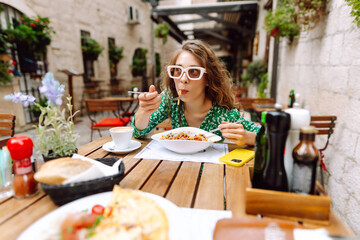 Beautiful tourist in bright green dress enjoying meal outdoors in cafe. Young woman having lunch in cafe in old town. Food and consumption concept, tourism.