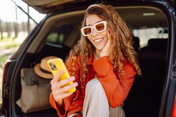 Cheerful female traveler with a phone sits in the trunk of a car and blogs or texts. Young woman...