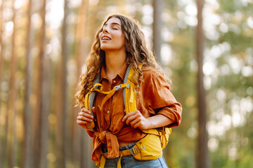Cheerful female traveler with a hiking backpack enjoying hiking at sunset. Beautiful woman with curly hair feels freedom and happiness in the forest. Adventure concept.