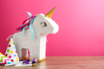 Beautiful pinata in shape of unicorn, party cones and blowers on wooden table against pink...