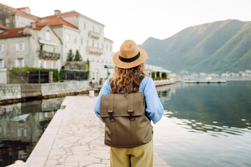 A rear view of a young tourist in a hat and with a backpack walking along the embankment. A...