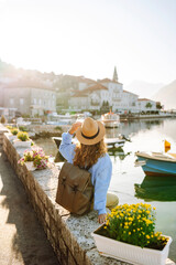 A rear view of a young tourist in a hat and with a backpack walking along the embankment. A...