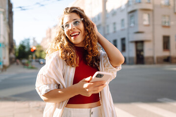 Fototapeta premium Cheerful woman with a phone walking on a sunny city street. Beautiful tourist chatting, blogging outdoors. Fashion concept. Lifestyle.