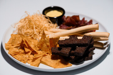 Assortment of savory snacks on a white plate including tortilla chips, cheese straws, bacon, breadsticks and creamy dip presented for enjoyment