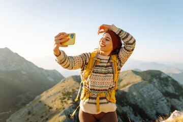 Hiker with bright hiking gear taking selfie on mountain. Cheerful woman feeling freedom and enjoying sunset on trail. Hiking, adventure concept. Active lifestyle.